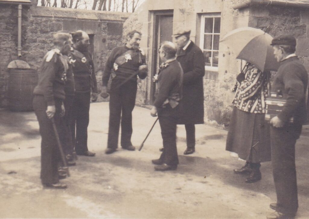 Cecil Sharp shown talking with five rapper sword dancers at Beadnell, Northumberland. On the right or the photograph is a Bessie, carrying a parasol, and a one-row melodeon player.